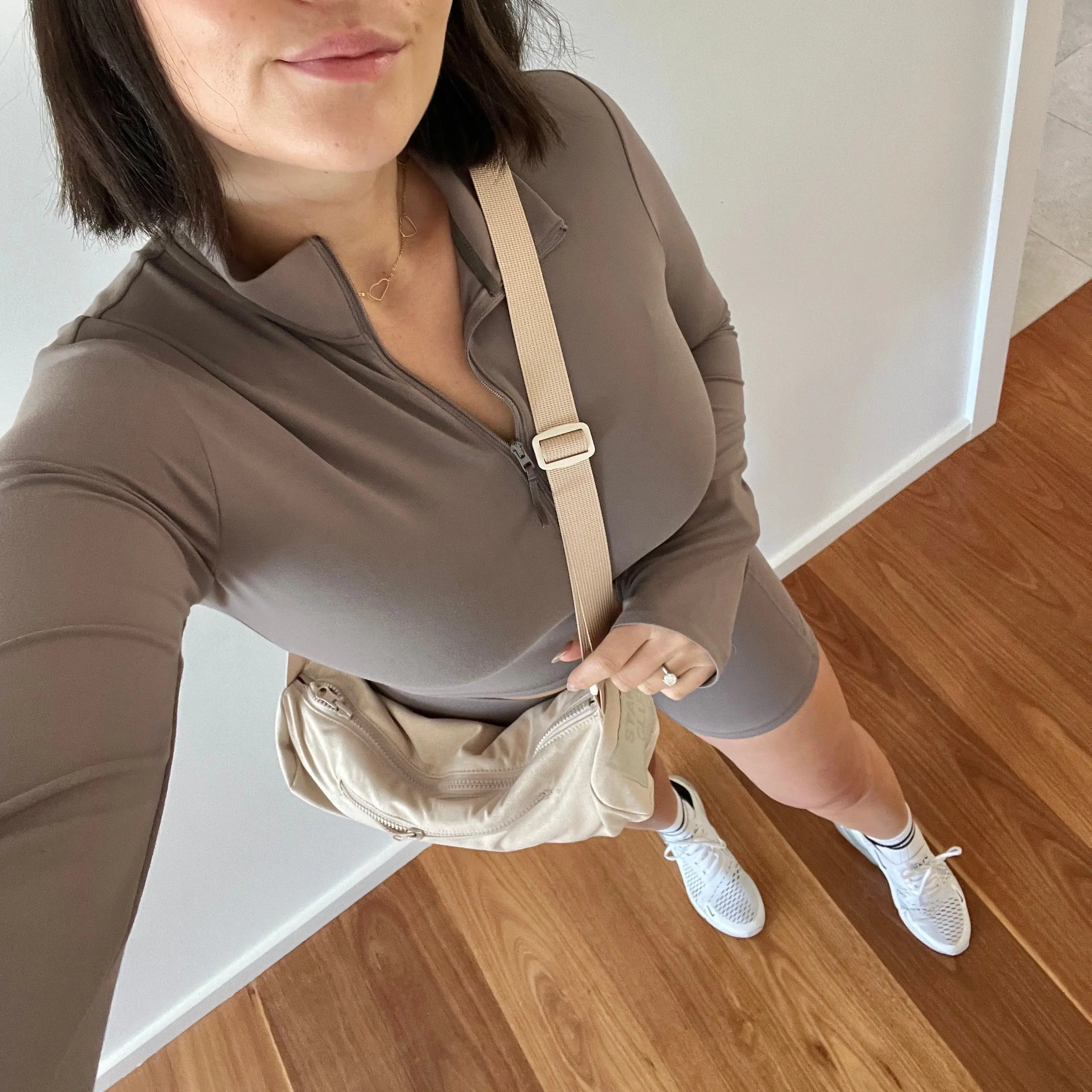 Woman in a brown shirt and shorts holding a beige crossbody bag indoors.
